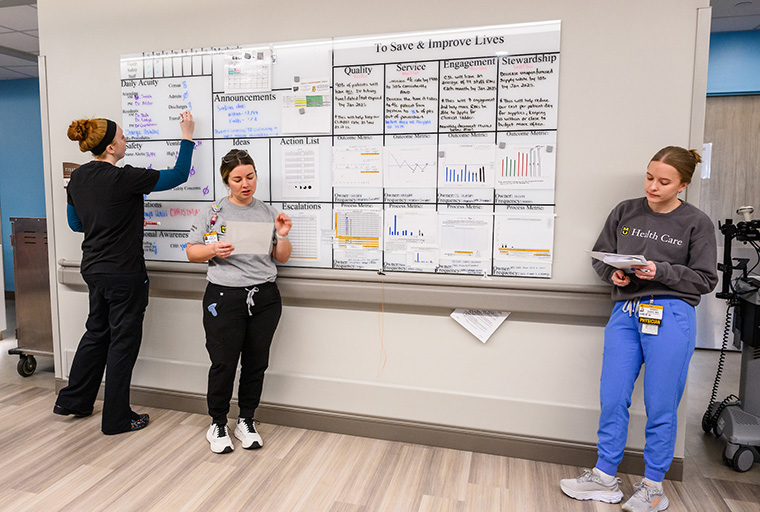  Nurse Ashley Hubbard is leading the Tier 1 Huddle in the pediatric intensive care unit with resident physician Dr. Sarah Shah following along and nurse Macy Walquist updating the huddle board.